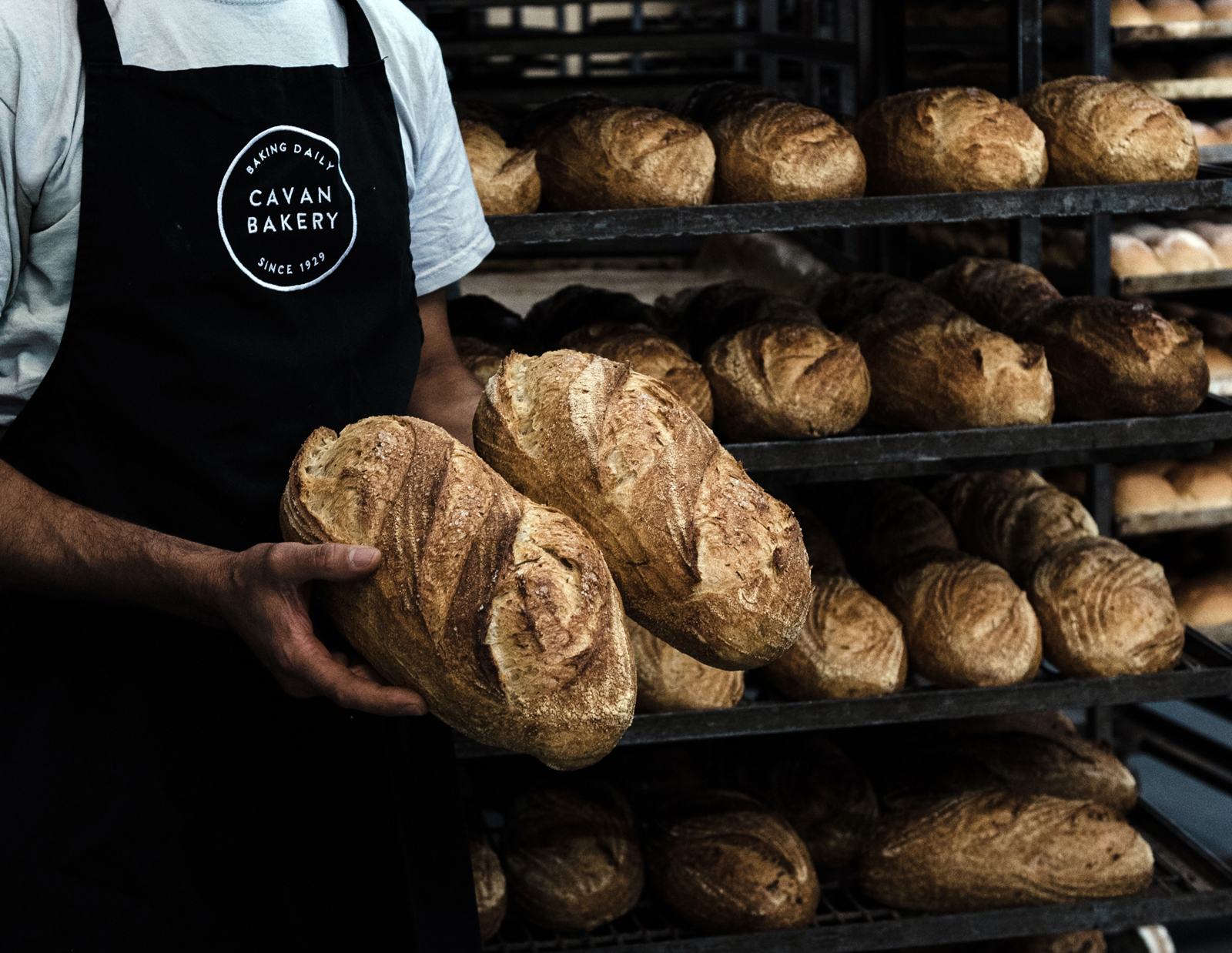 A baker in a Cavan Bakery apron holding two fresh loaves in front of the racks
